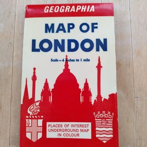 May include: A red and white box with the text "GEOGRAPHIA MAP OF LONDON" in blue and white. The box features a silhouette of St. Paul's Cathedral and other London landmarks. The text "Scale - 6 inches to 1 mile" is printed below the title. The box also includes the text "PLACES OF INTEREST UNDERGROUND MAP IN COLOUR".