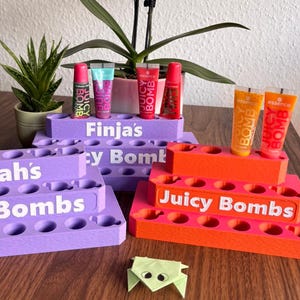 Könnte beinhalten: Drei abgestufte Holz-Organizer in Lila und Orange mit weißem Text, der Sarah's Juicy Bombs, Finja's Juicy Bombs und Juicy Bombs lautet. Jeder Organizer verfügt über mehrere kreisförmige Aussparungen zur Aufbewahrung von Essence Juicy Bomb Lipgloss-Tuben.