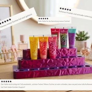 Könnte beinhalten: Ein mehrstufiger, magentafarbener Kosmetik-Organizer hält mehrere Tuben "Juicy Bomb" Lipgloss in verschiedenen Farben. Der Organizer hat ein geometrisches Design und wird auf einer Holzoberfläche ausgestellt.