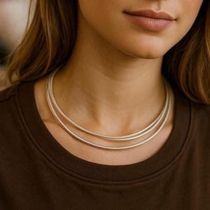 Puede incluir: Un conjunto de collares plateados con tres finas hebras redondas. Los collares se llevan alrededor del cuello, sobre una camiseta marrón. La joyería es sencilla y elegante.