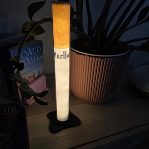 Puede incluir: Una lámpara cilíndrica con forma de cigarrillo que presenta una sección de filtro naranja brillante y un cuerpo blanco con el texto Marlboro. La lámpara se mantiene vertical sobre una base negra de forma irregular, emitiendo luz suave.