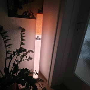 Könnte beinhalten: Eine hohe, zylindrische Stehlampe, die wie eine Zigarette gestaltet ist. Der obere Teil weist ein beiges, gesprenkeltes Filtermuster auf, während der untere Bereich weiß ist und den schwarzen Schriftzug 'Marlboro' vertikal an der Seite trägt.