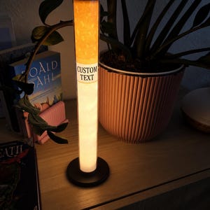 Op de afbeelding: Een hoge, cilindervormige lamp in de vorm van een sigaret, met een oplichtend oranje bovendeel en een wit onderste deel. De lamp heeft een zwarte ronde voet en toont de woorden CUSTOM TEXT in zwarte letters op een witte band in het midden.