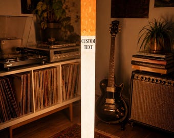 Peut inclure: Un lampadaire cylindrique haut en forme de cigarette, doté d'une section filtre orange lumineuse au sommet et d'un corps blanc avec les mots CUSTOM TEXT imprimés en noir. La lampe repose sur une base métallique sombre à quatre pieds.