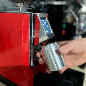 May include: A red and silver coffee grinder with a digital display. A hand is holding a silver portafilter under the grinder spout.