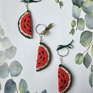 Könnte beinhalten: Drei gehäkelte Wassermelonen-Schlüsselanhänger mit grünen, weißen und roten Details. Jeder Schlüsselanhänger hat einen silbernen Ring und ein grünes Band.