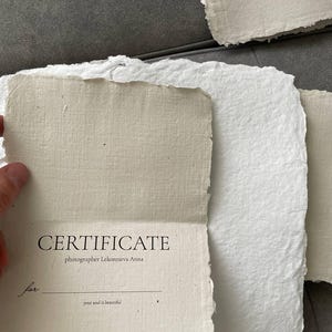May include: A stack of handmade paper sheets, some with deckled edges, in shades of white and beige. One sheet is open, revealing the word "CERTIFICATE" and the photographer's name. The text includes "Certificate valid 3 months".