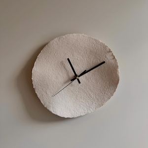 Puede incluir: Un reloj de pared redondo, de color blanquecino, con una superficie texturizada. Tiene manecillas negras y un diseño minimalista. El reloj está montado en una pared gris claro, creando una estética neutra y moderna. Diámetro: aprox. 25 cm.