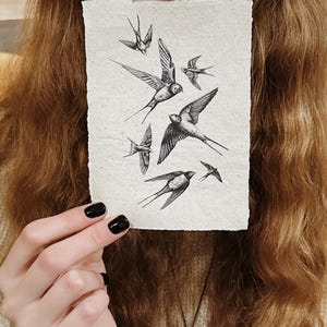 May include: A rectangular artwork featuring a flock of swallows in flight, rendered in black ink on textured, off-white paper. The birds are depicted with detailed wings and bodies, creating a dynamic composition. The artwork is held up in front of a person.