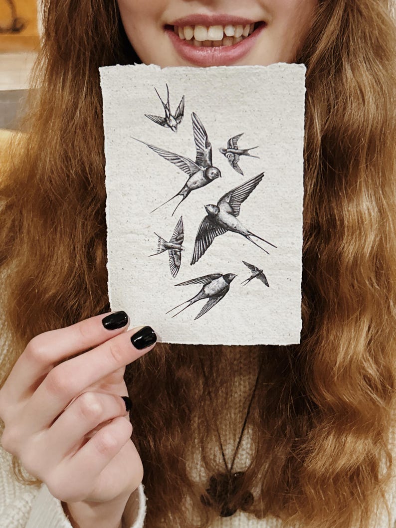May include: A rectangular artwork featuring a flock of swallows in flight, rendered in black ink on textured, off-white paper. The birds are depicted with detailed wings and bodies, creating a dynamic composition. The artwork is held up in front of a person.
