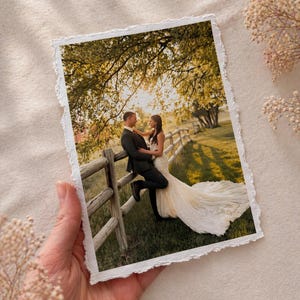 May include: A photograph with a torn edge shows a couple embracing near a wooden fence. The bride wears a white wedding dress with a long train. The groom is in a black suit. The background features a green field and trees.