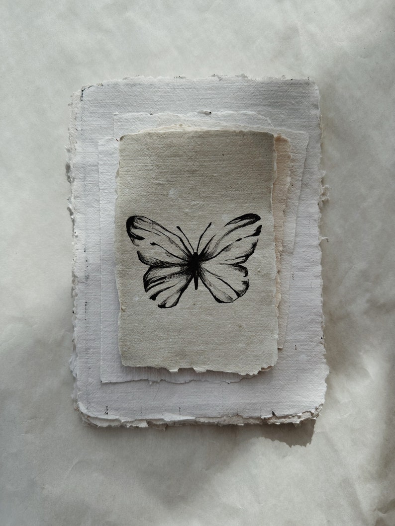 May include: A stack of handmade paper with a black ink butterfly illustration. The butterfly is centered on a smaller, light-colored paper, layered on top of larger, textured white sheets.