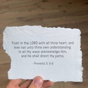 May include: A torn piece of white paper with the text of Proverbs 3:5-6 printed in black. The paper is held by a hand, and the background is a wooden surface.