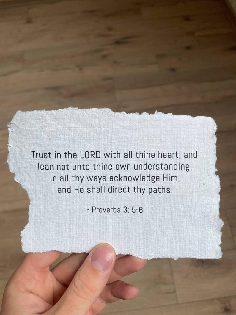 May include: A torn piece of white paper with the text of Proverbs 3:5-6 printed in black. The paper is held by a hand, and the background is a wooden surface.