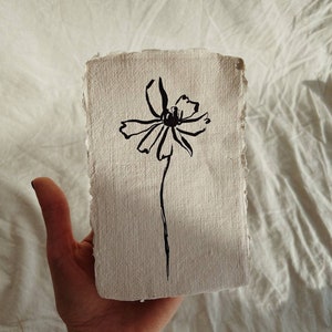 May include: A black ink drawing of a flower on a textured, off-white paper. The flower has delicate petals and a dark center, with a thin stem. The paper has a rough, torn edge, giving it a handmade appearance. The artwork is simple and elegant.