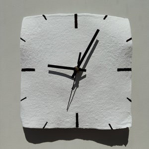Puede incluir: Reloj blanco cuadrado con manecillas y marcadores de hora negros. El reloj tiene una superficie texturizada y un centro color latón. Las manecillas proyectan una sombra sobre la esfera. El reloj está montado en una pared gris claro.