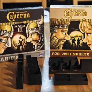 Puede incluir: Dos cajas de juego de mesa, "Caverna: Cueva contra Cueva", con ilustraciones de figuras tipo vikingo. La caja de la izquierda es negra con texto "EPOCHE II" y "ERWEITERU". La caja de la derecha es beige con texto "FÜR ZWEI SPIELER". Organizadores negros de componentes del juego están al frente.