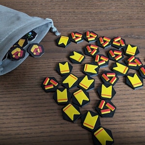 Puede incluir: Primer plano de una bolsa de terciopelo gris con piezas de juego hexagonales negras y amarillas que se derraman. Las piezas tienen un diseño amarillo y rojo.