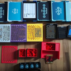 Könnte beinhalten: Eine Sammlung von Brettspielkomponenten, darunter Kartenboxen, perforierte Tabletts und kleine Behälter in verschiedenen Farben. Die Kartenboxen sind blau und schwarz, mit weißem Text und Zahnradgrafiken. Weitere Farben sind Weiß, Lila, Gelb, Schwarz, Rot und Rot-Orange.