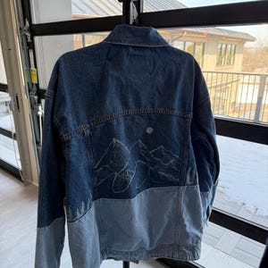 Könnte beinhalten: Zweifarbige Jeansjacke mit dunklerem Blau oben und hellblauem Abschnitt unten. Auf der Rückseite der Jacke befindet sich ein weißes Bergketten-Design. Sie hängt an einem schwarzen Kleiderbügel.