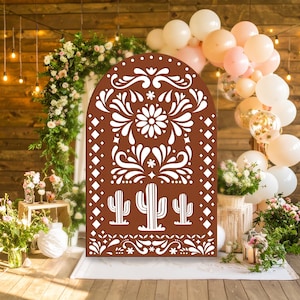 May include: A brown wooden arch with white floral and cactus cutouts, set against a backdrop of balloons, flowers, and string lights. The arch is the focal point, suggesting a festive or celebratory theme, possibly for a wedding or party.
