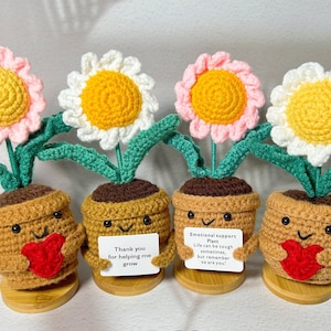 May include: Four handmade crochet flower pot decorations. Each pot has a smiling face, a red heart, and a sign with text. The flowers have yellow centers and pink or white petals. The pots are on small wooden bases.