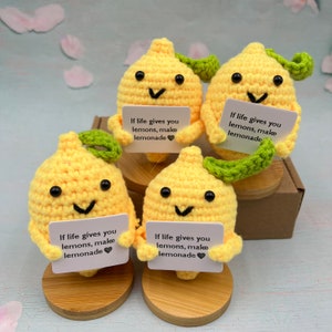 Pode incluir: Quatro figuras de limão de crochê amarelo feitas à mão com rostos sorridentes, olhos pretos e detalhes de folhas verdes. Cada uma segura uma pequena placa branca com o texto "If life gives you lemons, make lemonade". As figuras são exibidas em suportes de madeira.