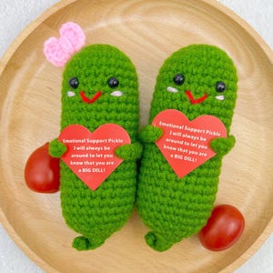 May include: Two green crocheted pickle plushies holding red heart-shaped cards with the text "Emotional Support Pickle I will always be around to let you know that you are a BIG DILL!"