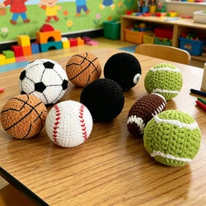 May include: A collection of handmade crochet sports balls, including a soccer ball, basketballs, a baseball, a football, a pool ball, and tennis balls. The balls are in various colors and sit on a wooden table.
