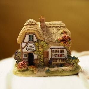 Könnte beinhalten: Miniaturfigur eines Cottage mit Strohdach, Schornstein und dem Schild "The Baker's Dozen". Das Cottage ist mit Fenstern, einer Tür und farbenfrohen floralen Akzenten versehen. Die Figur steht auf einem kleinen Sockel.