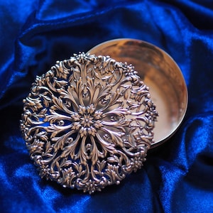 Op de afbeelding: Een zilverkleurig, rond compact poederdoosje met een open deksel. Het deksel heeft een ingewikkeld, bloemen filigraan ontwerp. Het compact poederdoosje ligt op een diepblauwe, getextureerde stof.