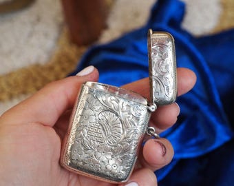 May include: A silver rectangular box with an open lid, featuring intricate floral and scrollwork engravings. The box is held in a hand, showcasing the detailed craftsmanship and antique design. The box is likely a vintage item.