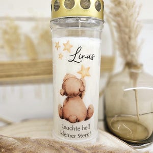 Könnte beinhalten: Eine zylindrische weiße Grabkerze in einem transparenten Kunststoffbehälter mit goldenem Deckel. Auf der Vorderseite befindet sich eine Illustration eines braunen Teddybären, der nach einem Stern greift, der Name Linus und der Text Leuchte hell kleiner Stern!