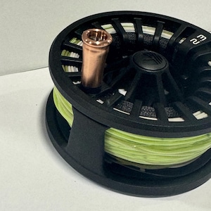 May include: A black fly fishing reel with a copper-colored arbor and bright green fly line. The reel has a modern design with a matte finish and is set against a neutral background. The reel is designed for fly fishing.