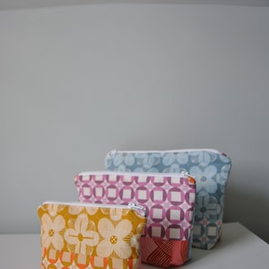 Könnte beinhalten: Drei Reißverschlusstaschen in verschiedenen Größen. Die größte Tasche ist blau mit einem weißen Blumenmuster. Die mittlere Tasche ist rosa mit einem weißen geometrischen Muster. Die kleinste Tasche ist gelb mit einem weißen Blumenmuster und einem geometrischen Muster am Boden.