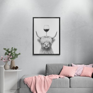 May include: Black and white framed print of a Highland cow with a wine glass balanced on its head. The artwork is displayed on a gray wall above a gray sofa with pink pillows and a pink throw blanket. A small white table with a vase of flowers is to the left.