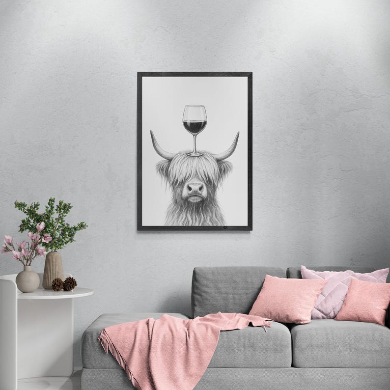 May include: Black and white framed print of a Highland cow with a wine glass balanced on its head. The artwork is displayed on a gray wall above a gray sofa with pink pillows and a pink throw blanket. A small white table with a vase of flowers is to the left.