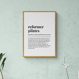 May include: A framed print with the words "reformer pilates" in a bold, black font. The definition of reformer pilates is below the title. The frame is gold, and the background is a light teal color.