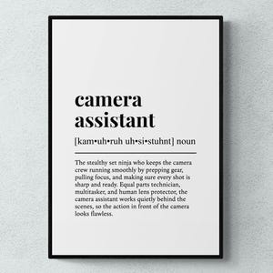 May include: Black framed print with the words "camera assistant" in bold, black font. Below, the phonetic pronunciation is given. The text describes the role of a camera assistant.