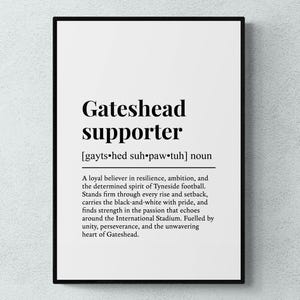 May include: A black framed print featuring the words "Gateshead supporter" in a bold, black font. The definition below describes a loyal believer in the spirit of Tyneside football. The print is displayed against a white wall.