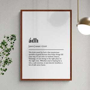 Op de afbeelding: Een ingelijste print met het Ierse woord "&aacute;dh" in zwarte tekst op een witte achtergrond. De definitie van het woord is ook opgenomen. De lijst is donkerbruin. De print hangt aan een lichtgrijze muur.