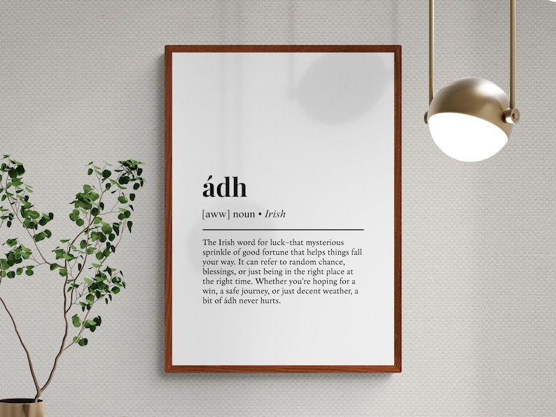 Op de afbeelding: Een ingelijste print met het Ierse woord "&aacute;dh" in zwarte tekst op een witte achtergrond. De definitie van het woord is ook opgenomen. De lijst is donkerbruin. De print hangt aan een lichtgrijze muur.