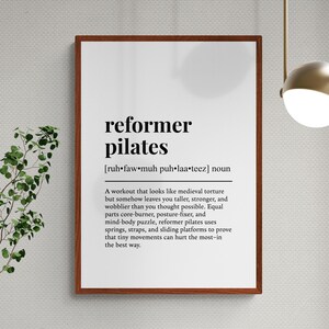 May include: A framed print with the words "reformer pilates" in bold black font. The definition below describes the workout. The print is in a brown frame and hangs on a textured white wall. A plant is to the left.