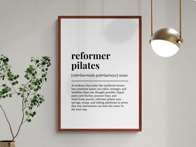 May include: A framed print with the words "reformer pilates" in bold black font. The definition below describes the workout. The print is in a brown frame and hangs on a textured white wall. A plant is to the left.