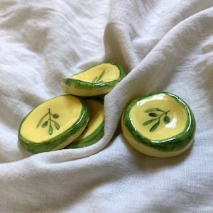 Può includere: Una collezione di piatti in ceramica fatti a mano a forma di oliva. Ogni piatto presenta un interno giallo con un disegno di ramo d'ulivo verde e un bordo verde. I piatti sono disposti su un tessuto bianco.