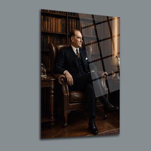 May include: A portrait of Mustafa Kemal Atatürk wearing a dark suit, white shirt, and tie, seated in a brown leather armchair. The background features a wooden bookshelf filled with numerous books and a globe on a side table in a library setting.