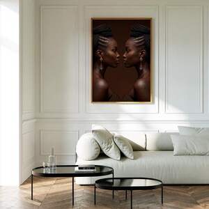 Puede incluir: Una impresión artística enmarcada que presenta a dos mujeres de perfil, mirándose. Tienen piel oscura, peinados trenzados y pendientes. La obra de arte, sobre un fondo marrón, está en un marco dorado, sobre un sofá blanco.