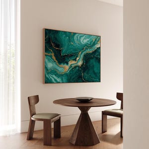 Puede incluir: Una gran obra de arte mural cuadrada con marco dorado presenta un diseño abstracto en tonos turquesa, verde esmeralda y dorado. Debajo, una mesa redonda de madera y dos sillas de madera con cojines verdes completan el comedor.