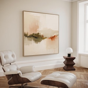Op de afbeelding: Een abstract schilderij in een houten lijst hangt aan een crèmekleurige muur. Het kunstwerk toont zachte penseelstreken in beige, perzik en olijfgroen. Een witte leren stoel en voetenbank met houten accenten staan op een parketvloer. Een kleine houten bijzettafel met een lamp is ook aanwezig.