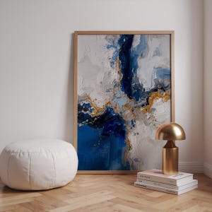 Könnte beinhalten: Abstrakter Kunstdruck in einem Holzrahmen mit strukturiertem Design in Blau, Weiß und Gold. Eine goldfarbene Tischlampe steht auf Büchern. Links befindet sich ein weißer, runder Hocker. Moderne Wohnkultur.